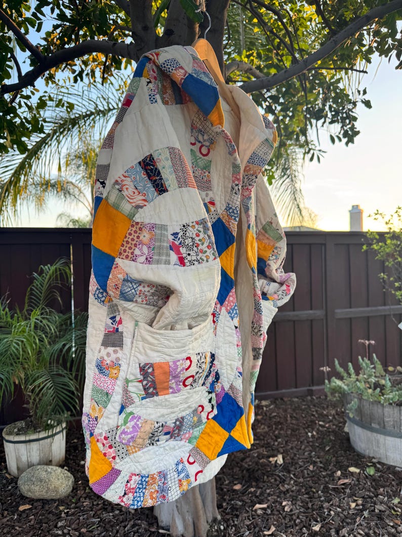 May include: A handmade quilt coat with a patchwork design. The coat features a variety of colorful fabric scraps in a mix of patterns, including floral, geometric, and solid colors. The coat is hanging on a wooden hanger outdoors.