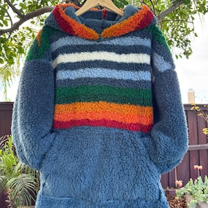 May include: A blue, hooded sweatshirt with a colorful striped design. The stripes include red, orange, green, white, and blue. The sweatshirt is made of a soft, textured material and has a kangaroo pocket.