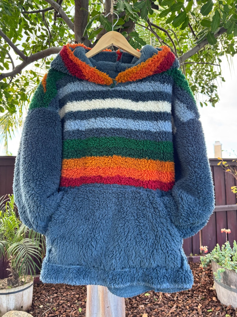 May include: A blue, hooded sweatshirt with a colorful striped design. The stripes include red, orange, green, white, and blue. The sweatshirt is made of a soft, textured material and has a kangaroo pocket.