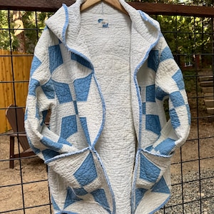 May include: A long-sleeved hooded jacket crafted from a white and light blue quilted fabric featuring a geometric star pattern. The garment has an open front, a hood, and two large patch pockets on the lower front, with blue trim along the edges.