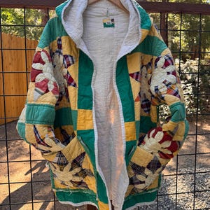 May include: A hooded jacket crafted from a patchwork quilt featuring a yellow, green, and white color palette with red and blue plaid accents. The garment displays a floral star pattern, a white interior lining, and two front pockets.