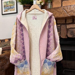 May include: A hooded jacket crafted from quilted fabric featuring a patchwork design in shades of pink, yellow, and blue. The garment has an open front, long sleeves, and two large patch pockets on the lower front. The interior is lined with cream fabric.
