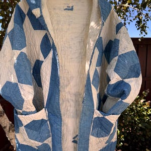 May include: A hooded jacket crafted from quilted fabric featuring a blue and white geometric patchwork pattern. The garment includes two front patch pockets, a light-colored interior lining, and a small rectangular label at the inner neckline.