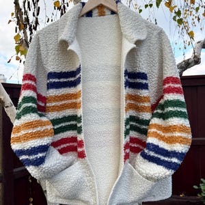 May include: A cream-colored fleece jacket with colorful horizontal stripes in red, blue, green, and orange on the sleeves. The jacket has a collared neckline, two front pockets, and is hanging on a wooden hanger.
