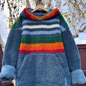 May include: A blue fleece hoodie with a rainbow stripe design across the chest. The hoodie has a kangaroo pocket and a hood with a colorful trim. The sleeves have blue cuffs. The hoodie is hanging on a wooden hanger.