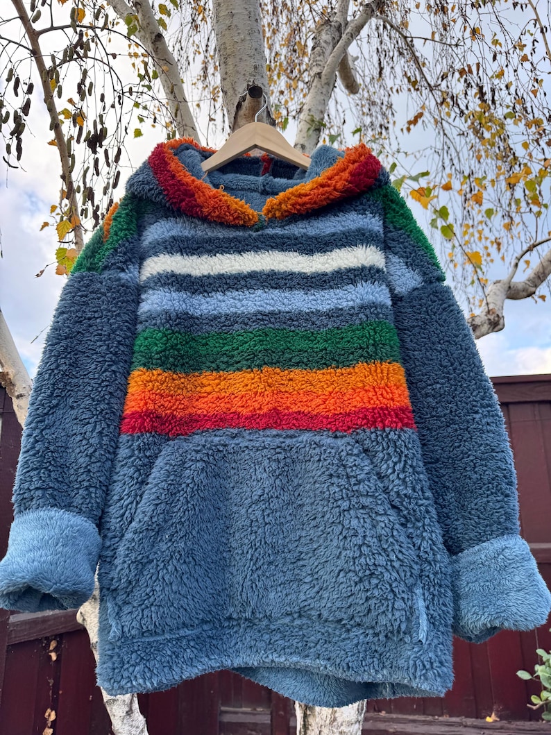 May include: A blue fleece hoodie with a rainbow stripe design across the chest. The hoodie has a kangaroo pocket and a hood with a colorful trim. The sleeves have blue cuffs. The hoodie is hanging on a wooden hanger.
