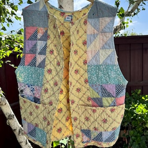 May include: A quilted vest with a patchwork design. The vest features a mix of floral, geometric, and solid fabric patterns in pastel colors, including yellow, blue, pink, and lavender. It has two front pockets and a yellow floral lining.