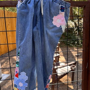 May include: Blue denim pants featuring an elastic waistband with a white drawstring. The pants are decorated with various fabric patches, including light pink and blue floral-shaped quilted appliqués and colorful plaid fabric inserts.