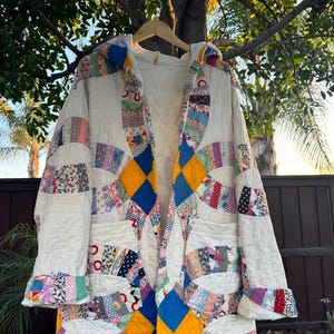 May include: A handmade, quilted coat with a cream-colored base and colorful patchwork details. The coat features a hood, pockets, and a diamond pattern in blue and yellow. The patchwork includes floral and geometric patterns.