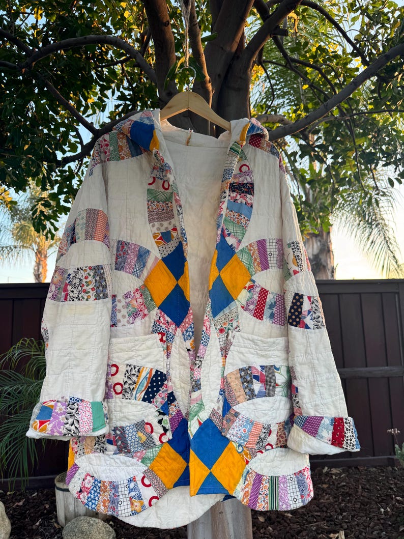 May include: A handmade, quilted coat with a cream-colored base and colorful patchwork details. The coat features a hood, pockets, and a diamond pattern in blue and yellow. The patchwork includes floral and geometric patterns.