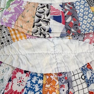 May include: Close-up of a vintage quilt with a variety of patterned fabric strips. The quilt features floral, geometric, and abstract designs in colors like purple, blue, orange, and white. The strips are sewn together with white fabric in between.