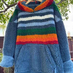 May include: A blue, hooded sweatshirt with a colorful striped design. The stripes include white, green, orange, and red. The sweatshirt has a kangaroo pocket and blue cuffs. The hood is trimmed with a fluffy, colorful material.