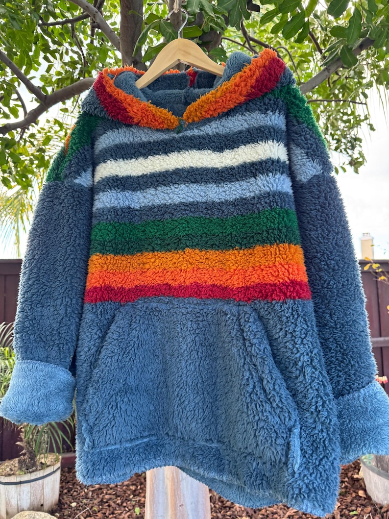 May include: A blue, hooded sweatshirt with a colorful striped design. The stripes include white, green, orange, and red. The sweatshirt has a kangaroo pocket and blue cuffs. The hood is trimmed with a fluffy, colorful material.