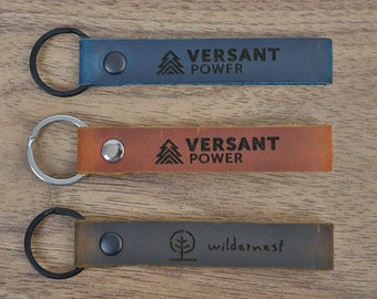 May include: Five leather keychains with different logos and colors. The keychains are on a wooden surface. The logos include "Untethered Ministries", "Cosley Zoo Conservation", "Versant Power", and "Wilderness".
