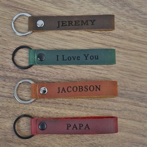 May include: Four leather keychains with metal rings. The keychains are brown, green, orange, and red. The keychains are engraved with the words "Jeremy", "I Love You", "Jacobson", and "Papa".
