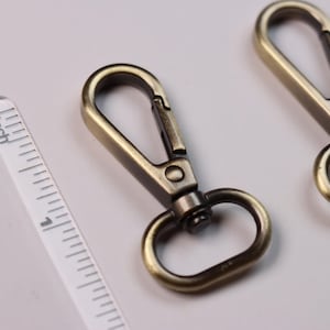 May include: Two antique brass-toned metal swivel lobster claw clasps with oval loops. A clear plastic ruler is positioned on the left side, showing the inch measurement markings along the edge of the hardware components.