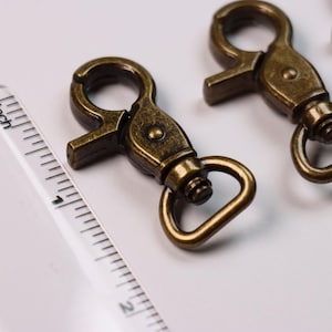 May include: Antique brass-colored metal swivel lobster claw clasps with a spring-loaded gate mechanism and a rotating D-ring base. A clear plastic ruler is positioned alongside the clasps, showing a length of approximately 1.5 inches.