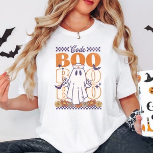 May include: White t-shirt with a graphic of a ghost wearing a nurse's hat and the text "Code Boo Boo" in orange and purple. The ghost is holding up two fingers in a peace sign.
