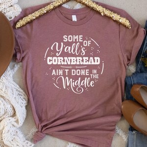 Peut inclure: Un t-shirt mauve avec le texte "Some of Y'all's Cornbread Ain't Done in the Middle" imprimé en blanc.