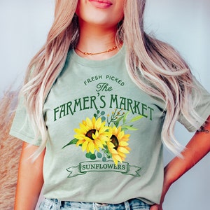 May include: A light green t-shirt with the text "Fresh Picked The Farmer's Market" in green lettering. The text is surrounded by two yellow sunflowers with green leaves and a banner that reads "Sunflowers".