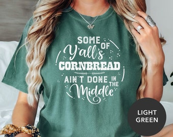 May include: A light green t-shirt with white text that reads "Some of Y'all's Cornbread Ain't Done in the Middle".