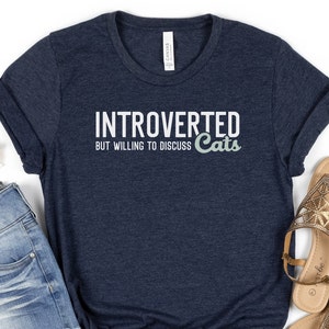 May include: A navy blue t-shirt with the text "Introverted but willing to discuss Cats" in white lettering. The word "Cats" is in a light green colour.