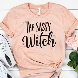 May include: A peach-colored t-shirt with the text "The Sassy Witch" in black lettering. The shirt has a distressed look and is decorated with small black stars.
