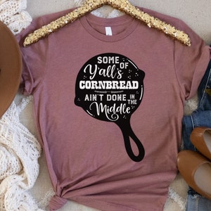 May include: A mauve t-shirt with a black graphic of a cast iron skillet. The text on the skillet reads "Some of Y'all's Cornbread Ain't Done in the Middle."
