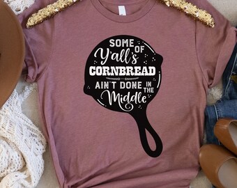 May include: A mauve t-shirt with a black graphic of a cast iron skillet. The text on the skillet reads "Some of Y'all's Cornbread Ain't Done in the Middle."