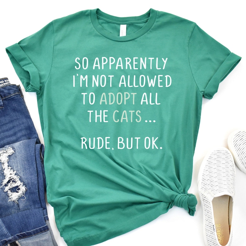 May include: A teal green t-shirt with the text "So apparently I'm not allowed to adopt all the cats... Rude, but ok." printed in white.