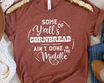 May include: A rust-colored t-shirt with white text that reads "Some of Y'all's Cornbread Ain't Done in the Middle".