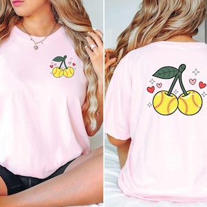 May include: Pink t-shirt with a graphic of two softball cherries with green stems and leaves. The cherries are yellow with red stitching. There are pink hearts and white stars around the cherries.