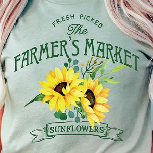 May include: A light blue t-shirt with the text "Fresh Picked The Farmer's Market Sunflowers" in green lettering. The text is surrounded by two yellow sunflowers with green leaves.