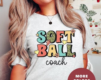 May include: A gray t-shirt with the text "Softball coach" in colorful, retro-style letters with a leopard print and other decorative elements. The text "More Colors Available" is in a red circle at the bottom of the image.