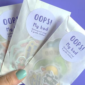 May include: Three white envelopes with a sticker on each. The stickers are round and white with purple text that reads "OOPS! My bad Waterproof vinyl B grade stickers".