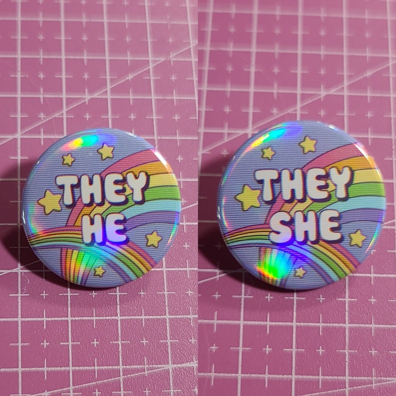 May include: Two iridescent rainbow pins with stars and the words "They He" and "They She" on a purple background.