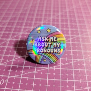 May include: A blue button with a rainbow and stars design. The text on the button reads "Ask me about my pronouns".