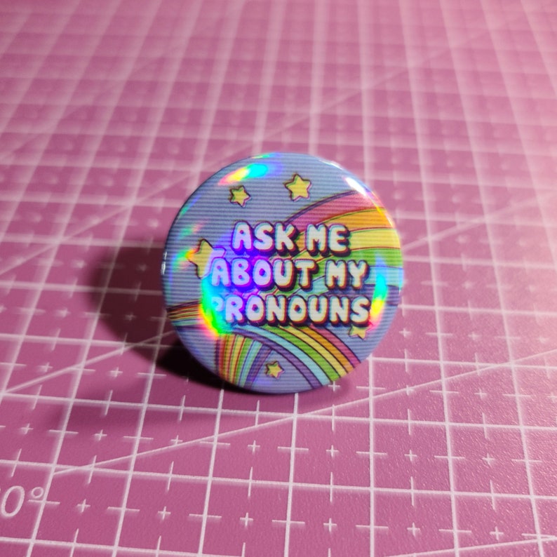 May include: A blue button with a rainbow and stars design. The text on the button reads "Ask me about my pronouns".