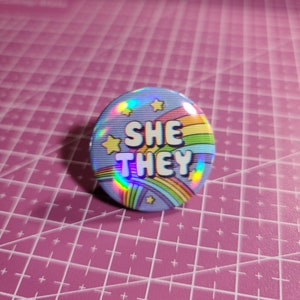 May include: A holographic button with a rainbow and stars design. The button says "SHE THEY" in bold, white letters.