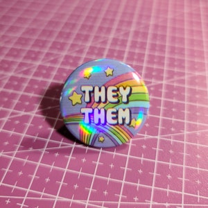May include: A blue button with a rainbow and stars design. The text "THEY THEM" is printed in white on the button.