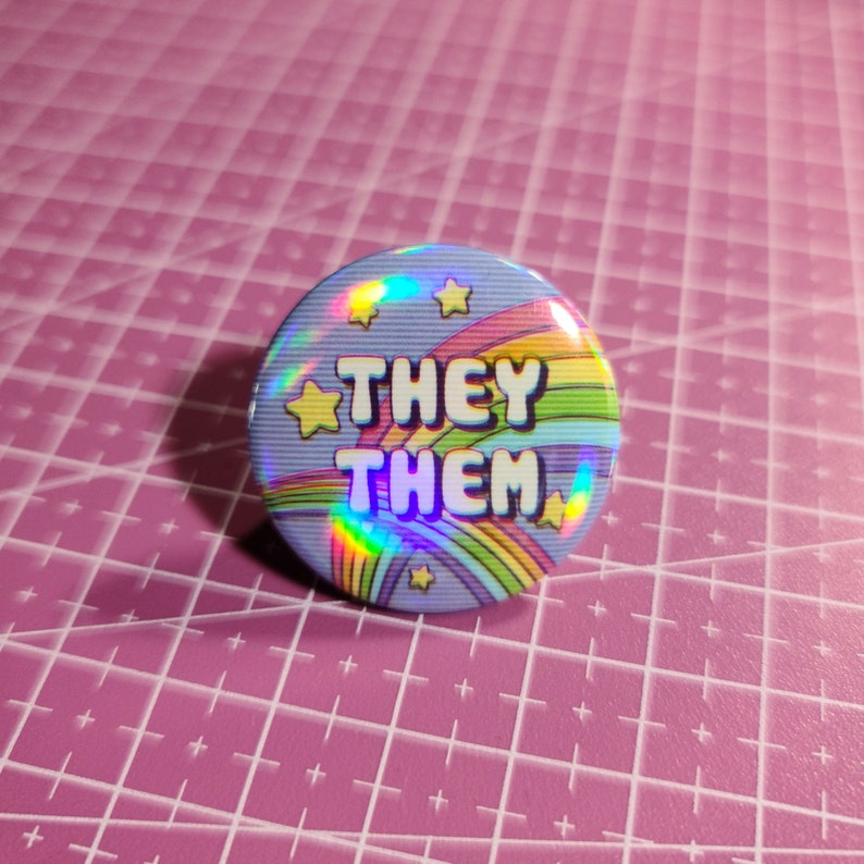 May include: A blue button with a rainbow and stars design. The text "THEY THEM" is printed in white on the button.