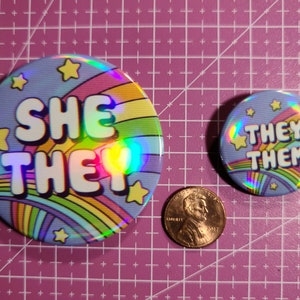 May include: Two round, holographic buttons with rainbow stripes and yellow stars. The larger button says "She They" and the smaller button says "They Them".