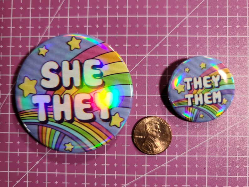 May include: Two round, holographic buttons with rainbow stripes and yellow stars. The larger button says "She They" and the smaller button says "They Them".