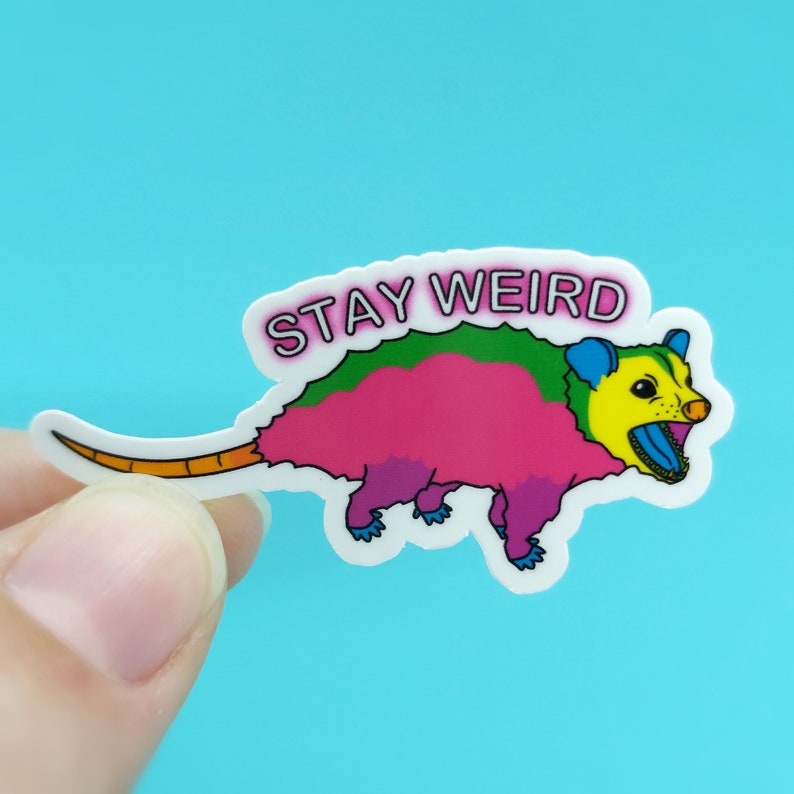 May include: A colorful sticker featuring a cartoon opossum with a pink body, green back, and yellow face. The sticker has the text "STAY WEIRD" in a white bubble. The opossum has a long orange tail and blue feet.