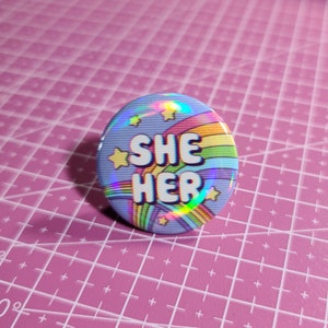 May include: A blue button with a rainbow and stars design. The text "SHE HER" is printed in white on the button.