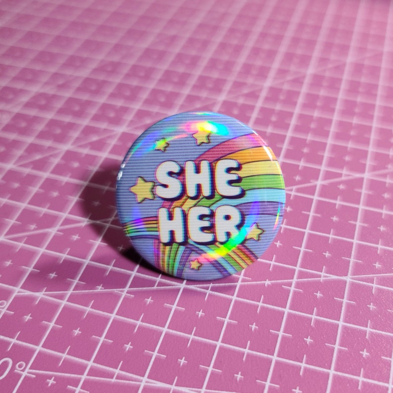 May include: A blue button with a rainbow and stars design. The text "SHE HER" is printed in white on the button.
