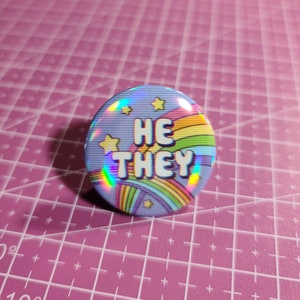 May include: A holographic button with a rainbow and stars design. The text "HE THEY" is printed in white on the button.