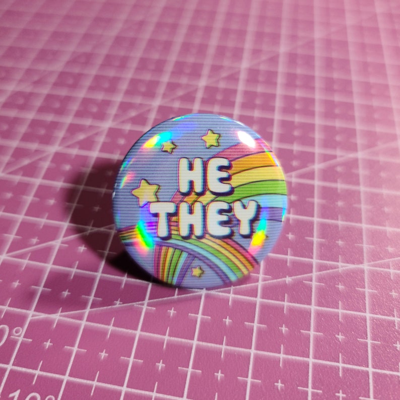May include: A holographic button with a rainbow and stars design. The text "HE THEY" is printed in white on the button.