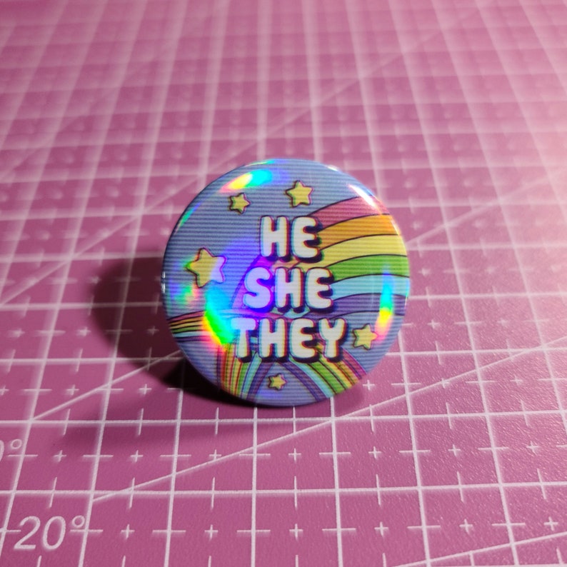May include: A blue button with a rainbow and stars design. The text "HE SHE THEY" is printed on the button.