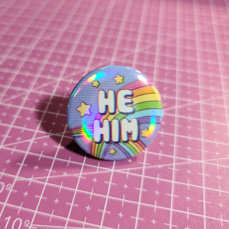 May include: A blue and rainbow holographic button with the text "HE HIM" and stars.
